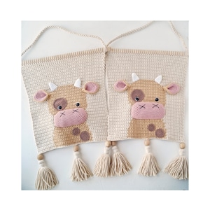 May include: Two cream-colored, crocheted wall hangings with cow faces. Each cow has a light brown face, pink nose and ears, and white horns. Wooden beads and cream tassels hang from the bottom. The hangings are connected by a cream-colored crocheted cord.