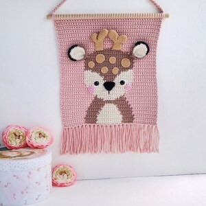 May include: A pink crocheted wall hanging with a brown deer face in the center. The deer has brown antlers, black eyes, and a white nose. The hanging has a fringe border.