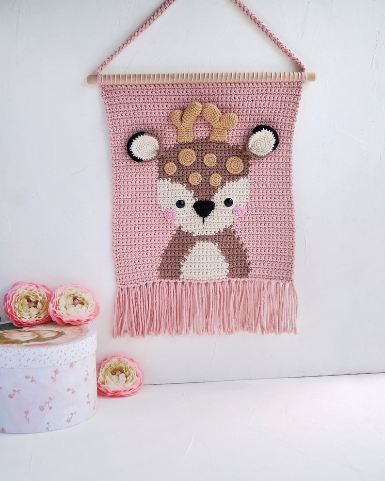 May include: A pink crocheted wall hanging with a brown deer face in the center. The deer has brown antlers, black eyes, and a white nose. The hanging has a fringe border.