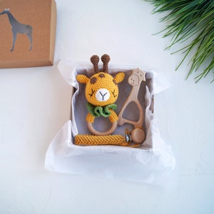 May include: A gift box filled with a crocheted giraffe rattle, a wooden giraffe teether, and a wooden teething ring. The box is lined with white tissue paper.
