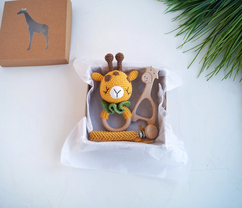 May include: A gift box filled with a crocheted giraffe rattle, a wooden giraffe teether, and a wooden teething ring. The box is lined with white tissue paper.