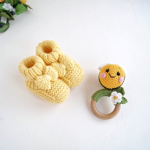 May include: A pair of yellow knitted baby booties with a bow detail and a crocheted bee rattle with a wooden ring. The bee has a smiling face with pink cheeks and a white flower.