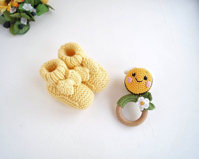 May include: A pair of yellow knitted baby booties with a bow detail and a crocheted bee rattle with a wooden ring. The bee has a smiling face with pink cheeks and a white flower.