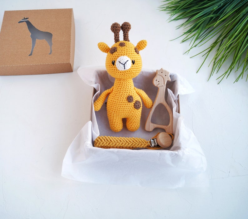 May include: A yellow crocheted giraffe toy with brown spots, a wooden giraffe teether, and a yellow crocheted teething necklace with a wooden clip in a white gift box.