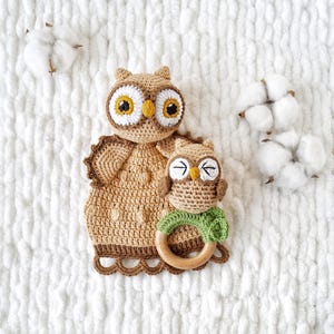 May include: Two crocheted owl toys in shades of brown, tan, and white, with large yellow eyes. One is a larger owl with a scalloped edge, and the other is a smaller owl holding a wooden ring with a green leaf. The owls are on a white, textured surface.