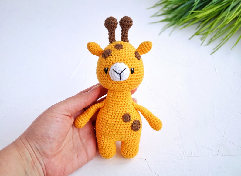 May include: A crocheted yellow giraffe toy with brown spots and ears. The giraffe has a white face with black eyes and a small white nose.