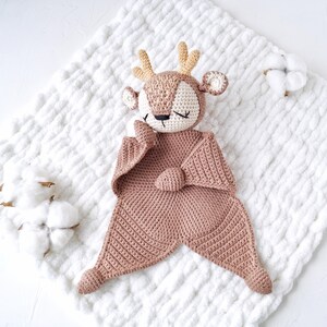 Peut inclure: Une couverture de s&eacute;curit&eacute; en crochet en forme de cerf, de tons beige et cr&egrave;me, avec des yeux ferm&eacute;s et de petits bois. La couverture pr&eacute;sente un motif g&eacute;om&eacute;trique textur&eacute; et repose sur une surface blanche matelass&eacute;e. Des brins de coton sont en arri&egrave;re-plan.