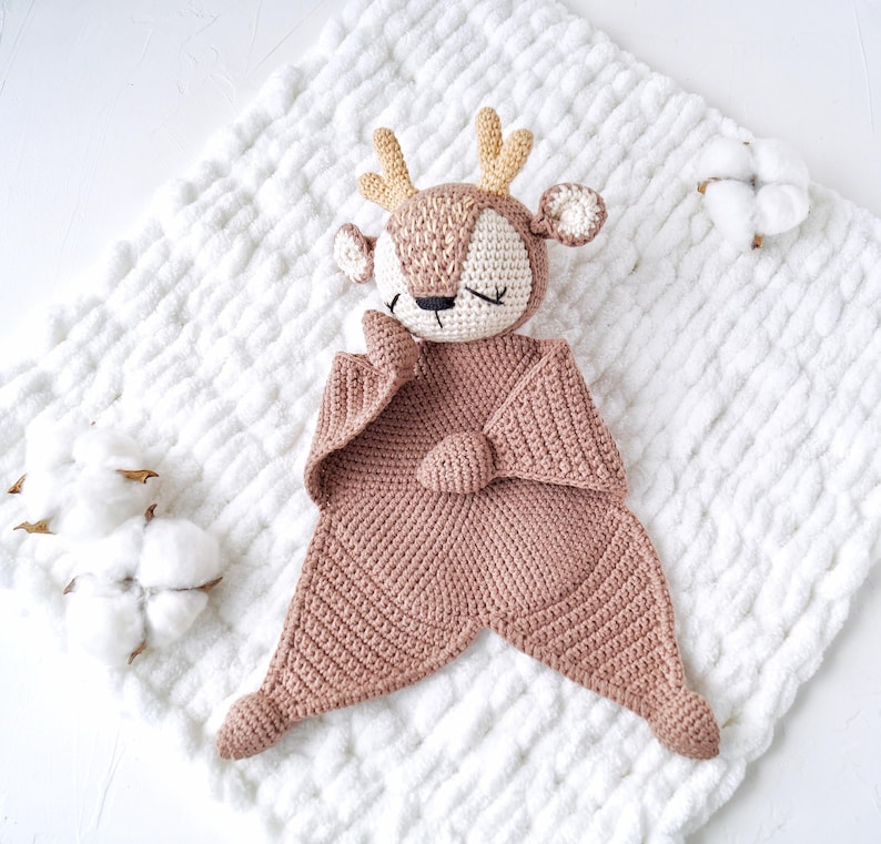Peut inclure: Une couverture de s&eacute;curit&eacute; en crochet en forme de cerf, de tons beige et cr&egrave;me, avec des yeux ferm&eacute;s et de petits bois. La couverture pr&eacute;sente un motif g&eacute;om&eacute;trique textur&eacute; et repose sur une surface blanche matelass&eacute;e. Des brins de coton sont en arri&egrave;re-plan.