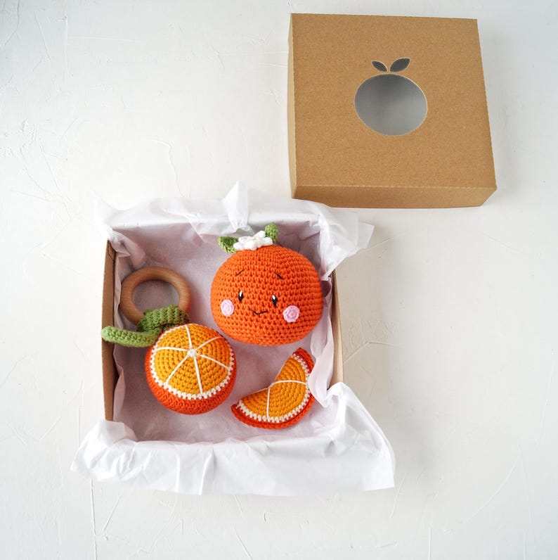 May include: A collection of orange-themed crochet baby toys, including a rattle, a round toy with a face, and a slice. The toys are presented in a cardboard box with a circular aperture and white tissue paper. The toys are orange, white, and green.