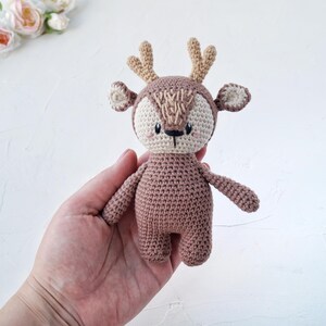 Peut inclure: Un jouet de cerf crochet&eacute; &agrave; la main, dans des tons de marron, beige et beige clair. Le cerf a un visage d&eacute;taill&eacute; avec de petits yeux noirs et un nez cousu. Les bois et les oreilles sont de couleur beige clair. Le jouet est tenu dans une main.
