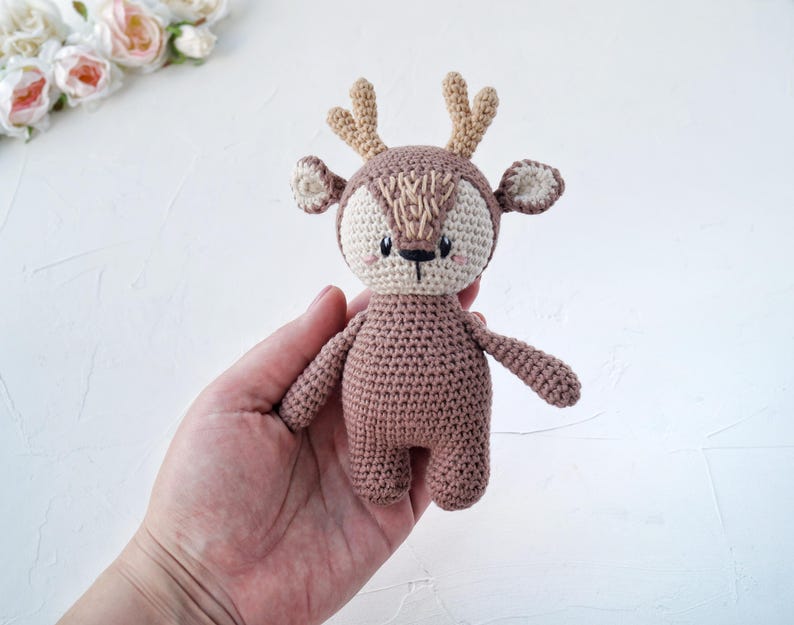 Peut inclure: Un jouet de cerf crochet&eacute; &agrave; la main, dans des tons de marron, beige et beige clair. Le cerf a un visage d&eacute;taill&eacute; avec de petits yeux noirs et un nez cousu. Les bois et les oreilles sont de couleur beige clair. Le jouet est tenu dans une main.