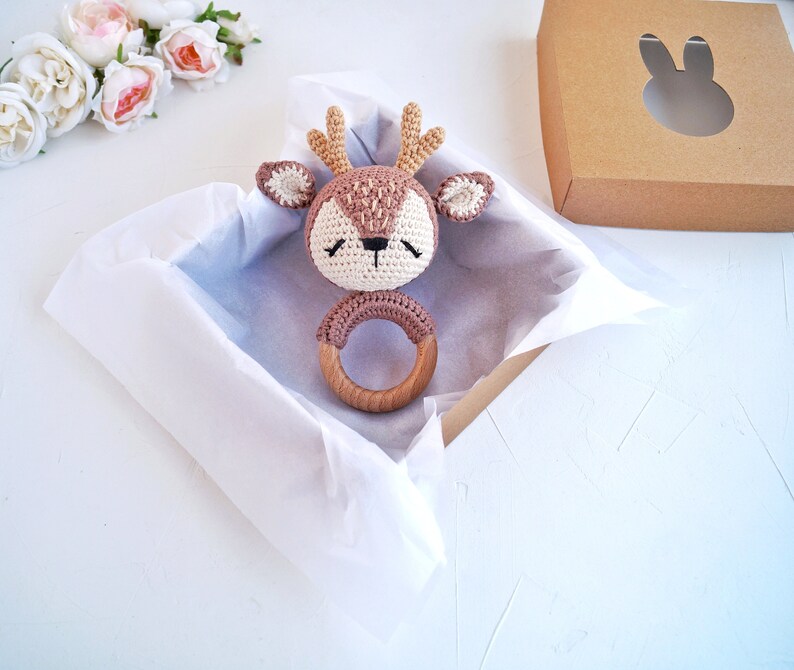 K&ouml;nnte beinhalten: Ein braunes und wei&szlig;es geh&auml;keltes Hirschr&auml;tsel mit einem Holzring. Das Rassel ist in einer wei&szlig;en, mit Seidenpapier ausgekleideten Geschenkbox.