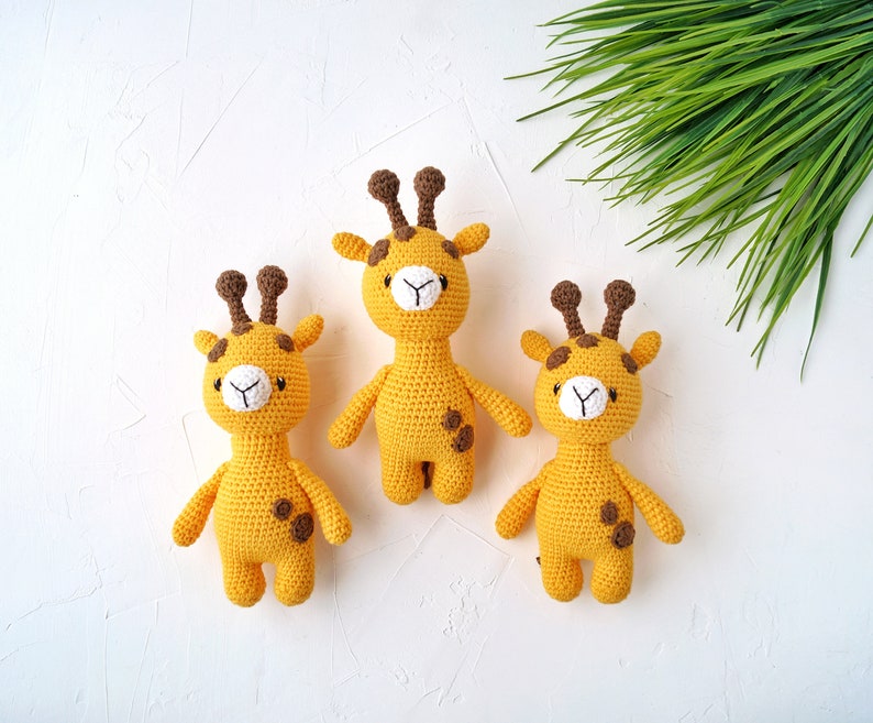 May include: Three crocheted giraffe toys with brown spots and white faces. The giraffes are standing on a white surface.