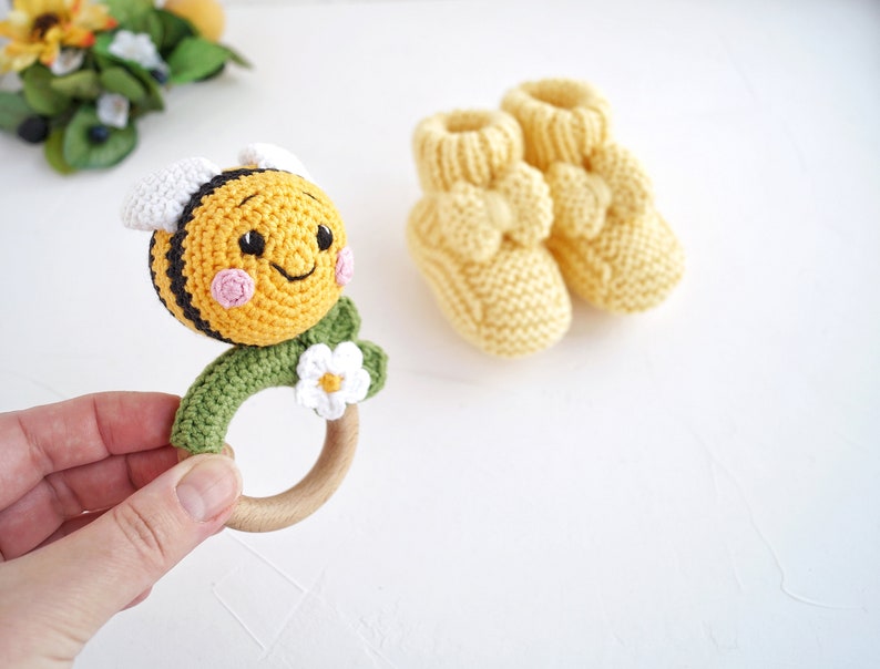 May include: A crocheted bee rattle with a wooden ring and a white flower. The bee is yellow with black stripes and has a smiling face.