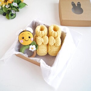 May include: A set of yellow knitted baby booties with a yellow and black bee rattle in a gift box. The booties have a bow on the top and the rattle has a flower on the side.