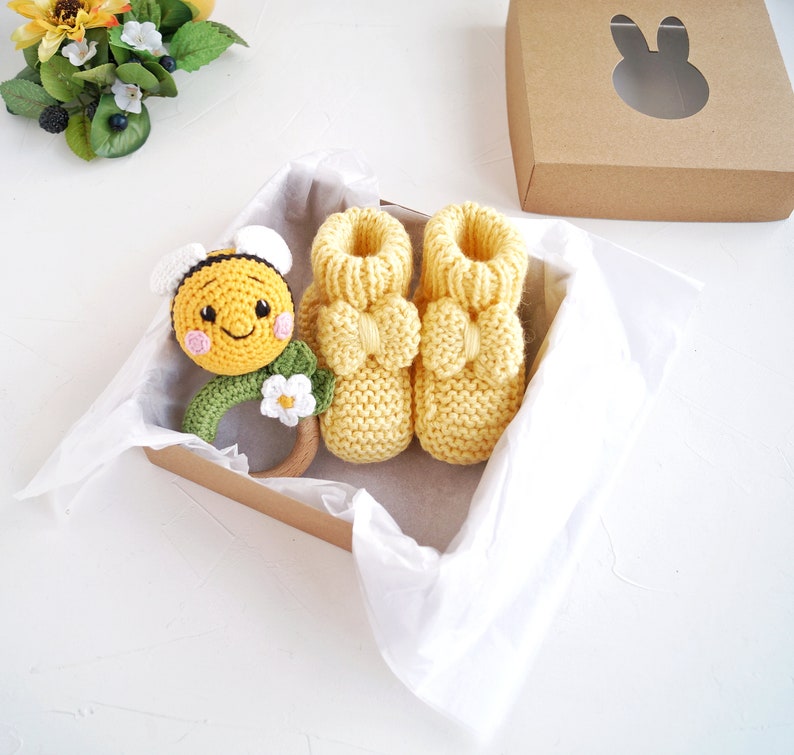 May include: A set of yellow knitted baby booties with a yellow and black bee rattle in a gift box. The booties have a bow on the top and the rattle has a flower on the side.