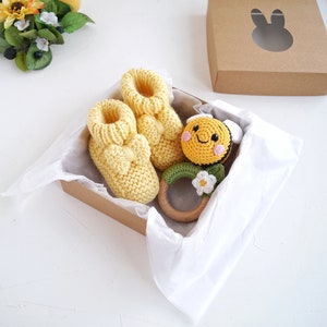 May include: A yellow gift box with white tissue paper lining. The box contains a pair of yellow knitted baby booties, a wooden teether with a crocheted bee and flower, and a brown gift box with a bunny cutout.
