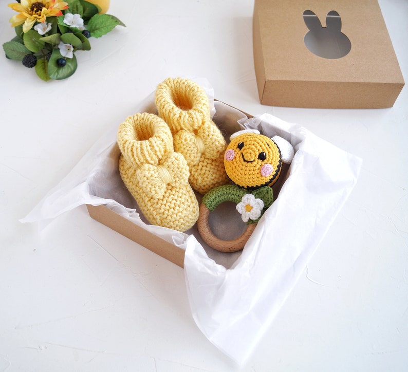 May include: A yellow gift box with white tissue paper lining. The box contains a pair of yellow knitted baby booties, a wooden teether with a crocheted bee and flower, and a brown gift box with a bunny cutout.