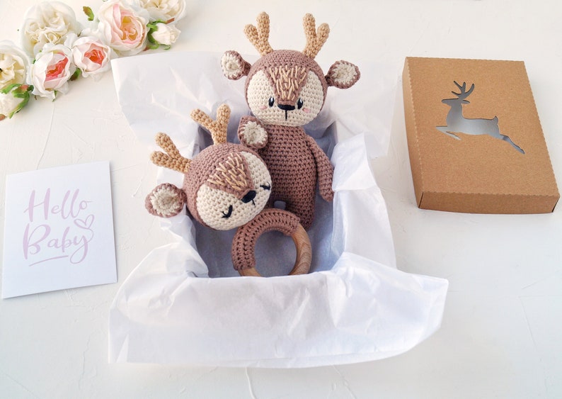 K&ouml;nnte beinhalten: Eine Geschenkbox gef&uuml;llt mit wei&szlig;em Seidenpapier enth&auml;lt zwei geh&auml;kelte Hirschspielzeuge. Ein Hirsch ist gr&ouml;&szlig;er und hat einen Holzring befestigt. Die Box ist braun mit einer ausgeschnittenen Hirschform. Eine Karte mit dem Text "Hello Baby" befindet sich im Hintergrund.