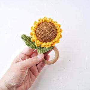 May include: A crocheted sunflower rattle toy with a wooden ring. The sunflower is yellow and brown with green leaves.