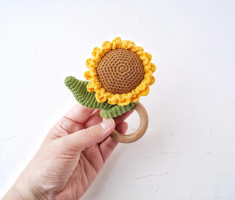 May include: A crocheted sunflower rattle toy with a wooden ring. The sunflower is yellow and brown with green leaves.