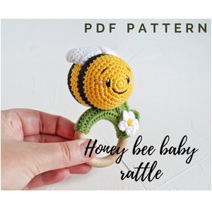 May include: A crocheted honey bee baby rattle with a wooden ring. The bee is yellow and black with a white face and a green leaf body. The rattle is held in a hand.