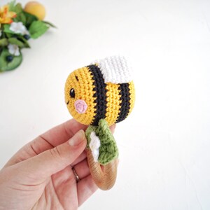 May include: A crocheted bee rattle toy with a wooden handle. The bee is yellow and black with a white wing and a pink face. The handle is decorated with green leaves and white flowers.