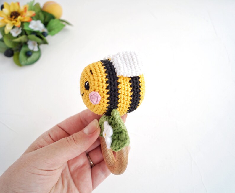 May include: A crocheted bee rattle toy with a wooden handle. The bee is yellow and black with a white wing and a pink face. The handle is decorated with green leaves and white flowers.
