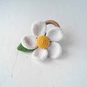 May include: A white crocheted flower with a yellow center and green leaves attached to a wooden ring. This is a baby teether.