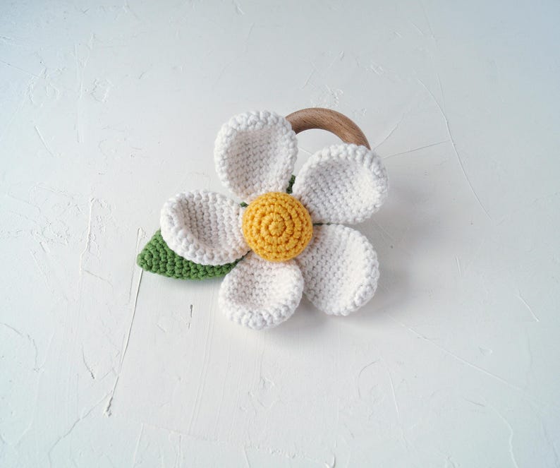 May include: A white crocheted flower with a yellow center and green leaves attached to a wooden ring. This is a baby teether.