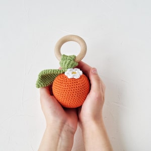 May include: A handmade crochet baby rattle in the shape of a fruit, featuring an orange body, green leaves, and a white flower. It includes a wooden ring for grasping. The toy is suitable for babies and toddlers.