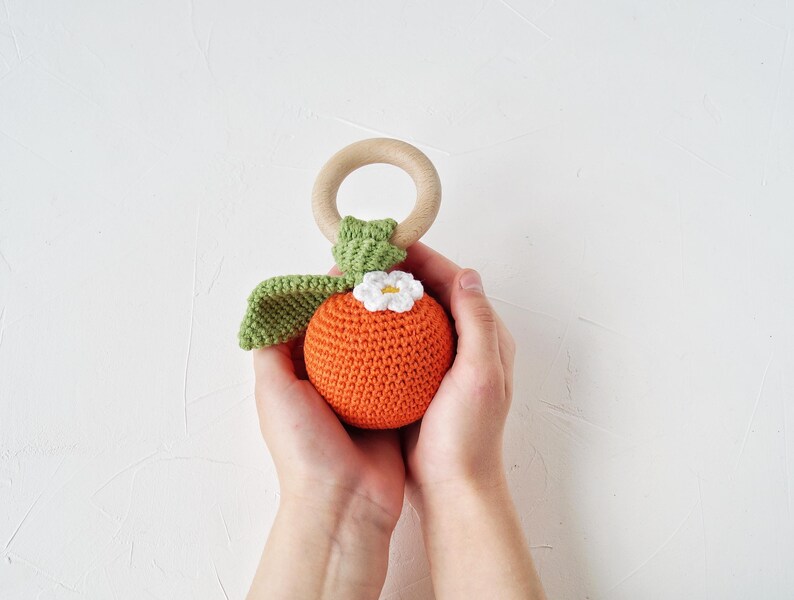 May include: A handmade crochet baby rattle in the shape of a fruit, featuring an orange body, green leaves, and a white flower. It includes a wooden ring for grasping. The toy is suitable for babies and toddlers.