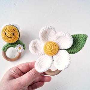 May include: Two crocheted baby teethers, one shaped like a daisy with a yellow center and green leaves, and the other shaped like a smiling sun with a yellow face and white eyes. Both teethers are attached to wooden rings.