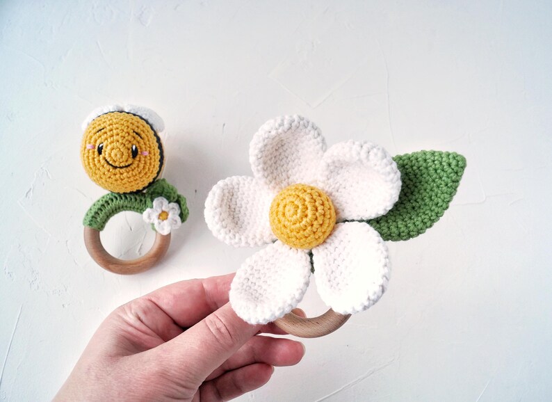 May include: Two crocheted baby teethers, one shaped like a daisy with a yellow center and green leaves, and the other shaped like a smiling sun with a yellow face and white eyes. Both teethers are attached to wooden rings.