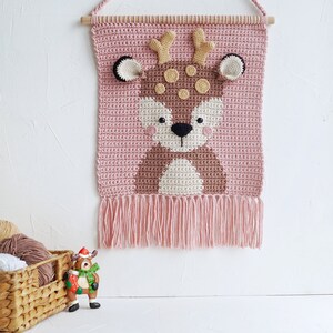 May include: A pink crocheted wall hanging with a brown deer face in the center. The deer has brown antlers, brown eyes, a white nose, and brown spots. The wall hanging has a fringe at the bottom.