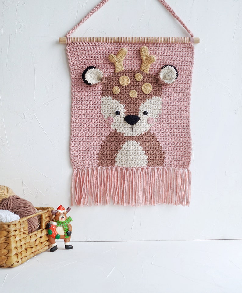 May include: A pink crocheted wall hanging with a brown deer face in the center. The deer has brown antlers, brown eyes, a white nose, and brown spots. The wall hanging has a fringe at the bottom.