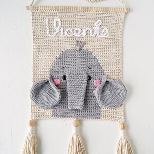 May include: A crocheted wall hanging with a gray elephant face and the name "Vicente" in white yarn. The hanging has two beige tassels.