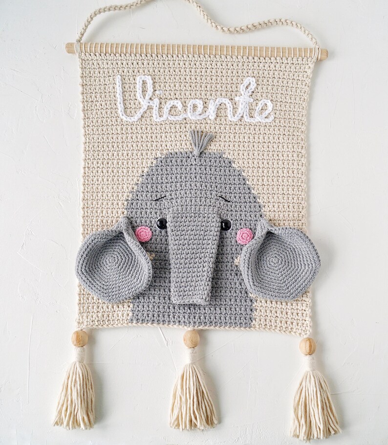 May include: A crocheted wall hanging with a gray elephant face and the name "Vicente" in white yarn. The hanging has two beige tassels.