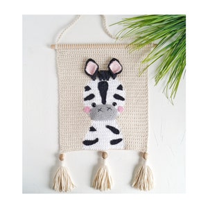 Op de afbeelding: Een beige gehaakte wandhanger met een zwart-wit zebra-ontwerp. De zebra heeft een grijze neus en roze wangen. De hanger heeft drie beige kwastjes.