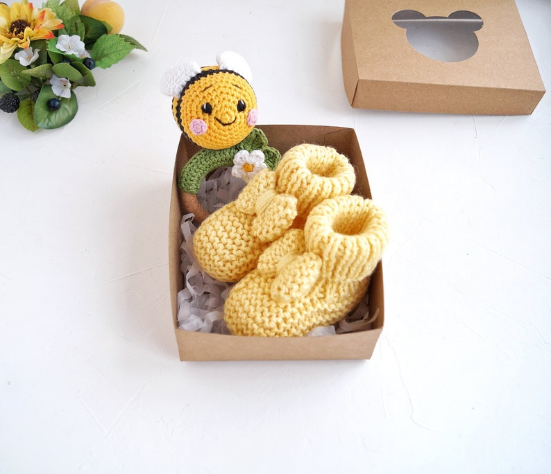 May include: A set of yellow knitted baby booties with a crocheted bee rattle in a brown gift box. The bee rattle has a green leaf and a white flower.