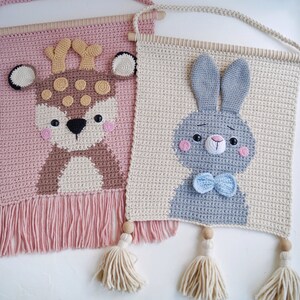May include: Two crocheted wall hangings with animal designs. The left one is pink with a brown deer with white spots. The right one is beige with a gray bunny with a blue bow tie.