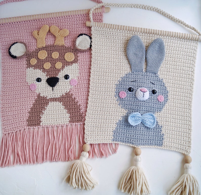 May include: Two crocheted wall hangings with animal designs. The left one is pink with a brown deer with white spots. The right one is beige with a gray bunny with a blue bow tie.
