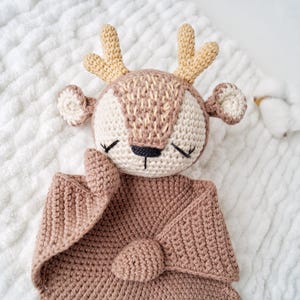 Peut inclure: Une couverture de s&eacute;curit&eacute; en crochet en forme de cerf, dans des tons beige et marron. Le cerf a un visage d&eacute;taill&eacute; avec les yeux ferm&eacute;s, de petites oreilles et des bois. La couverture est en mati&egrave;re douce et textur&eacute;e, pour b&eacute;b&eacute; ou enfant.