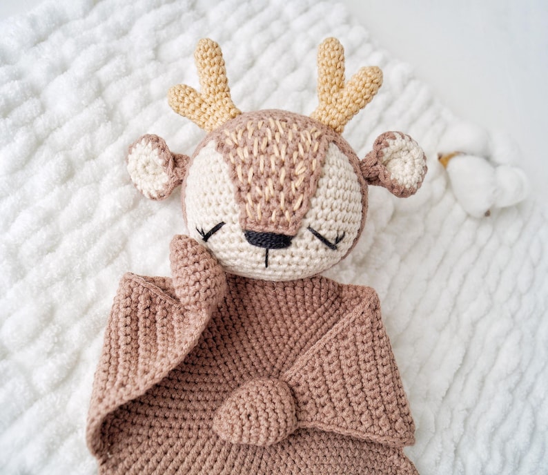 Peut inclure: Une couverture de s&eacute;curit&eacute; en crochet en forme de cerf, dans des tons beige et marron. Le cerf a un visage d&eacute;taill&eacute; avec les yeux ferm&eacute;s, de petites oreilles et des bois. La couverture est en mati&egrave;re douce et textur&eacute;e, pour b&eacute;b&eacute; ou enfant.