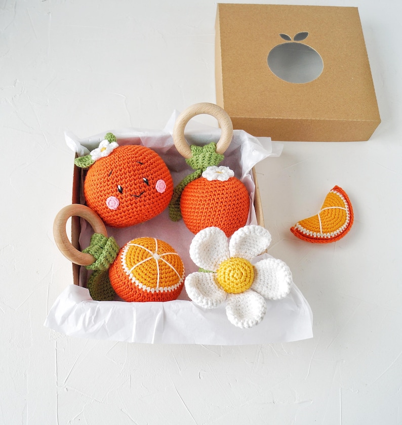 May include: A collection of orange crochet baby toys, including rattles and a flower, presented in a cardboard box. The toys feature wooden rings and white accents. The box has a die-cut orange shape on the lid.