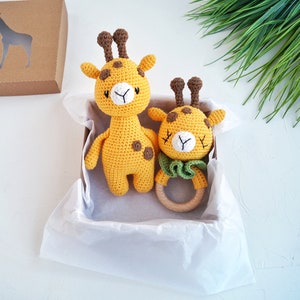 May include: A gift box filled with tissue paper contains two crocheted giraffe toys. One giraffe is larger and has brown spots, while the smaller giraffe is a rattle with a wooden ring.