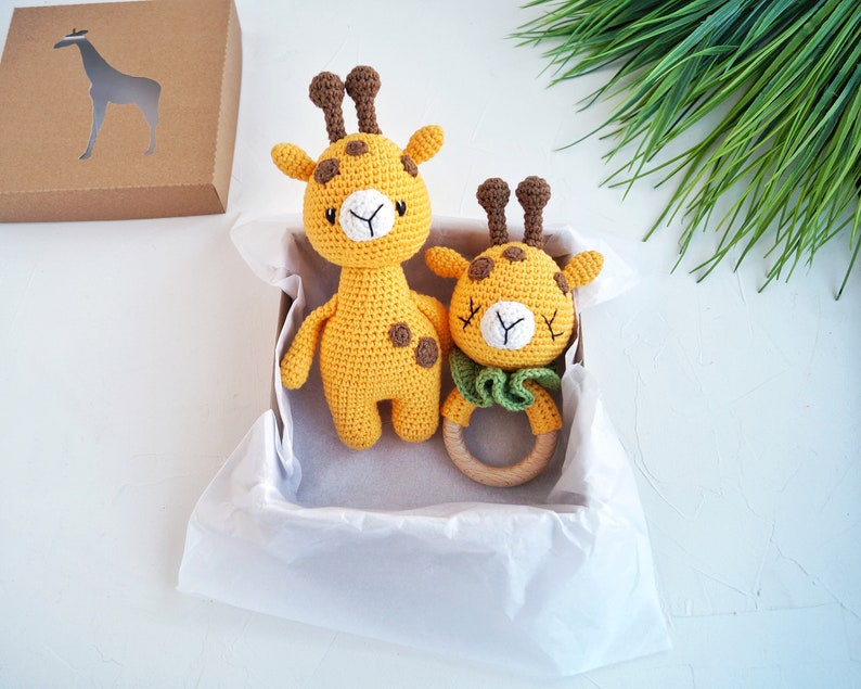 May include: A gift box filled with tissue paper contains two crocheted giraffe toys. One giraffe is larger and has brown spots, while the smaller giraffe is a rattle with a wooden ring.