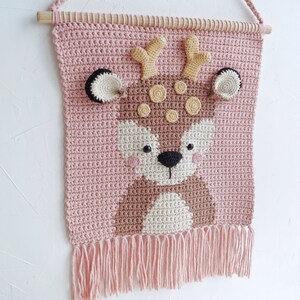 May include: A pink crocheted wall hanging with a brown deer face in the center. The deer has yellow antlers, brown eyes, and a black nose. The wall hanging has a fringe at the bottom.