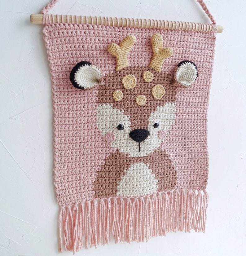 May include: A pink crocheted wall hanging with a brown deer face in the center. The deer has yellow antlers, brown eyes, and a black nose. The wall hanging has a fringe at the bottom.