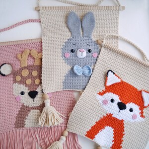 May include: Three crocheted wall hangings featuring animal designs. The first is a pink and brown deer with brown spots, the second is a beige bunny with a blue bow tie, and the third is a beige fox with orange and white fur.